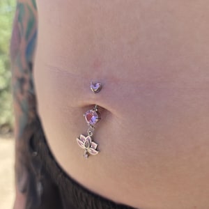 Puede incluir: Un piercing de ombligo plateado con una pequeña gema en forma de corazón en la parte superior, una piedra redonda de color rosa claro en el centro y un dije colgante de flor de loto con esmalte rosa claro y un pequeño cristal.