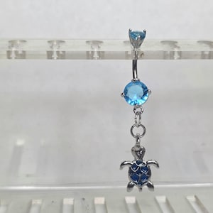 May include: A silver belly button ring with a light blue gem at the top and a larger round blue gem below. A small silver turtle charm with blue enamel detailing hangs from the bottom. The jewelry is displayed against a white background.