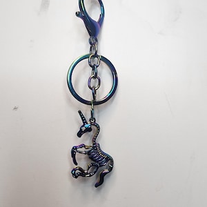 May include: A rainbow iridescent metal keychain featuring a skeleton unicorn charm. The keychain has a lobster clasp and a circular ring.