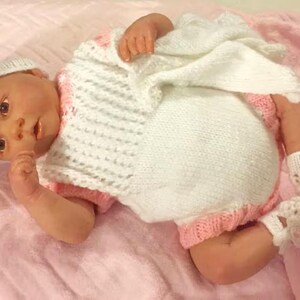 May include: A baby doll wearing a white and pink knitted outfit with a pink crocheted hat. The doll has realistic features and is lying on a pink blanket.