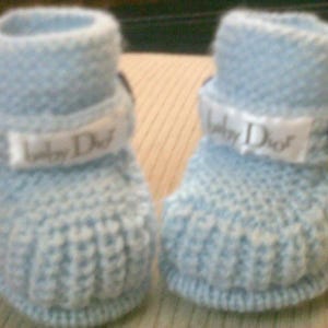 Könnte beinhalten: Ein Paar hellblaue gestrickte Babyschuhe mit weißem Band und dem Text "Baby Dior" auf jedem Schuh. Die Schuhe haben blaue Knöpfe an den Seiten.