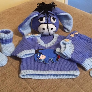 May include: A knitted baby outfit featuring a blue and light blue donkey character. The outfit includes a hat, sweater, trousers, booties, and a tail. The sweater has a donkey applique on the front.