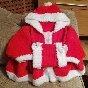 May include: Red knitted Santa suit with white trim and a hood. The suit has a button closure and white bows on the sleeves.