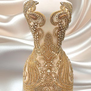 May include: A mannequin torso displays a couture garment. The dress is adorned with intricate gold beading and sparkling embellishments. The design features a sheer, nude-colored base with ornate patterns, creating a luxurious and detailed aesthetic. The background is a soft, flowing fabric.