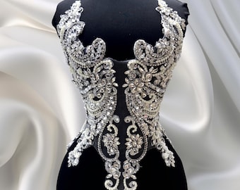 May include: A black dress form with a detailed bodice. The bodice is embellished with intricate silver and clear crystal beadwork. The design features floral and swirling patterns, creating a glamorous and elegant look. The dress form is set against a white satin backdrop.