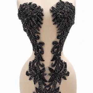 May include: A black beaded bodice applique on a mannequin. The intricate design features floral and leaf patterns, with the beads densely covering the sheer fabric. The applique is designed to be sewn onto a garment.