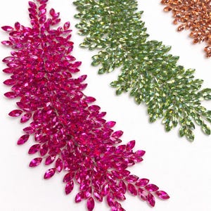 May include: Close-up of three decorative crystal leaf-shaped embellishments. The first is a vibrant fuchsia, the second is a light green, and the third is a copper color. Each is composed of many small, sparkling crystals.