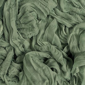 May include: Close-up of sage green fabric with a textured, wrinkled appearance. The fabric is draped and folded, creating a layered effect. The color is a muted, earthy green, and the material appears soft and lightweight.