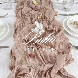May include: A dusty rose-colored, ruffled table runner with the word "Boho" in script. The runner is decorated with dried flowers and placed on a white tablecloth with place settings. The text "TABLE DECOR" and "DESIGNED FOR MOMENTS THAT MATTER" are also visible.
