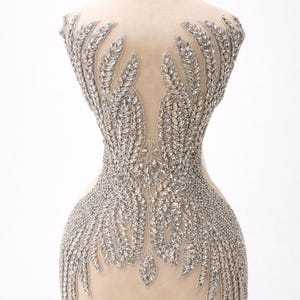 May include: A beige dress form adorned with an intricate silver crystal design. The embellishment features a symmetrical pattern of leaf-like shapes and sparkling crystals, creating a glamorous and elegant aesthetic. The form is set against a white background.