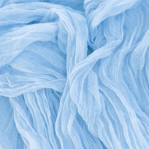 May include: A close-up shot of light blue fabric with a crinkled texture. The fabric is draped and folded, creating a flowing, wavy pattern. The soft material has a delicate, airy appearance, suggesting it could be used for clothing or decorative purposes.