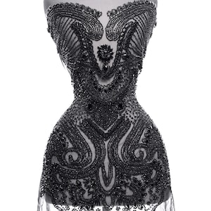 May include: A black beaded bodice on a mannequin. The bodice is intricately decorated with a variety of bead sizes and patterns. The design includes floral and swirling motifs. A sheer, black fabric extends from the bottom of the bodice.