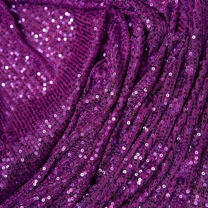 May include: Close-up of a vibrant purple fabric covered in small, reflective sequins. The fabric is draped, creating folds and shadows that highlight the texture and sparkle of the sequins. The overall effect is luxurious and eye-catching.