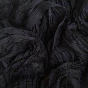 May include: Close-up of a dark charcoal-colored fabric with a crinkled texture. The fabric is draped and folded, creating a layered effect. The material appears to be lightweight and flowing, with visible wrinkles and folds.