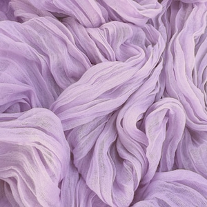 May include: Close-up of a light purple fabric with a textured, gauzy appearance. The fabric is draped and folded, creating soft waves and shadows. The color is a pale lavender, with variations in tone due to the folds.