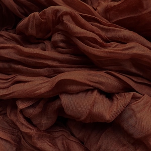 May include: Close-up of a flowing, textured fabric in a rich, warm brown hue. The material is draped and gathered, creating soft folds and shadows. The fabric appears lightweight and possibly sheer, with a subtle woven pattern.