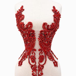 May include: A red beaded bodice with an intricate floral design. The bodice is embellished with numerous red gemstones, creating a sparkling effect. The design is symmetrical, with detailed patterns and a transparent center.