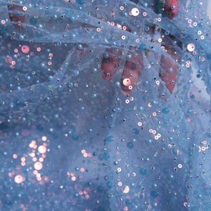May include: Close-up of light blue fabric with sequins and beads. The fabric is a sheer mesh, adorned with various sizes of iridescent sequins and small, round beads. The sequins and beads are in shades of blue, silver, and pink, creating a shimmering effect.