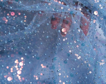 May include: Close-up of light blue fabric with sequins and beads. The fabric is a sheer mesh, adorned with various sizes of iridescent sequins and small, round beads. The sequins and beads are in shades of blue, silver, and pink, creating a shimmering effect.