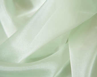 May include: A close-up of soft, light green fabric with a smooth, silky texture. The fabric is draped and folded, creating a flowing and elegant look.