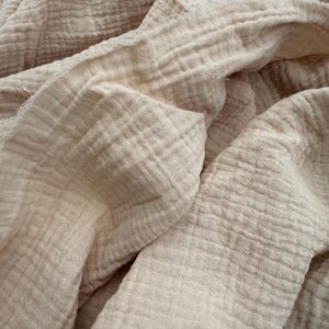 May include: Close-up of a soft, beige-colored fabric with a textured, crinkled appearance. The fabric is folded and draped, revealing the layers and the subtle variations in the material. The overall look is simple and natural.