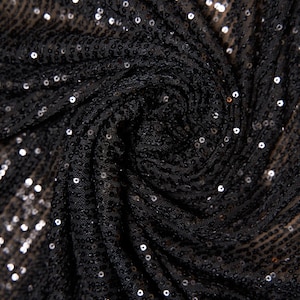 May include: Close-up of black sequin fabric. The fabric is draped and folded, with numerous small, shiny sequins scattered across the surface. The sequins catch the light, creating a sparkling effect. The fabric appears to be a dark, solid color.