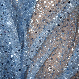 Puede incluir: Primer plano de una tela azul claro cubierta de lentejuelas pequeñas y redondas. Las lentejuelas son de tonos plateados y azules, creando un efecto brillante. La tela está drapeada, mostrando pliegues y textura, adecuada para ropa o manualidades.