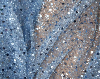 May include: Close-up of a light blue fabric covered in small, round sequins. The sequins are in shades of silver and blue, creating a shimmering effect. The fabric is draped, showing folds and texture, suitable for apparel or crafts.