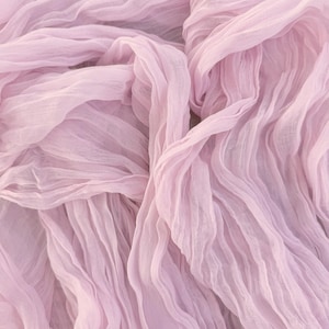 May include: A close-up shot of a light pink, flowing fabric. The material is gathered and draped, creating soft folds and a textured appearance. The fabric appears to be lightweight and semi-sheer, with a delicate, airy quality.
