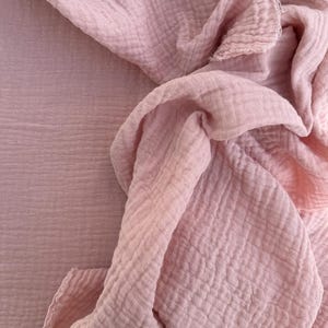 May include: A close-up of a soft, light pink fabric with a textured, crinkled appearance. The fabric is draped and folded, creating shadows and highlighting the material's texture. The overall aesthetic is delicate and airy.