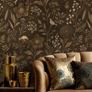 Puede incluir: Un papel pintado marrón oscuro con un patrón botánico de flores, trigo, pájaros y mariposas en tonos dorados y bronce. Un sofá de terciopelo beige se sitúa delante, adornado con un cojín floral dorado y un cojín oscuro estampado.