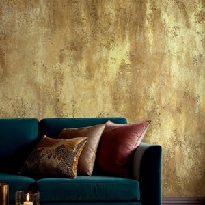 Modern living room interior with a teal velvet sofa and elegant pillows. Faux Gold Texture Wallpaper featuring a distressed metallic effect and rustic grunge finish for a sophisticated wabi sabi decor.