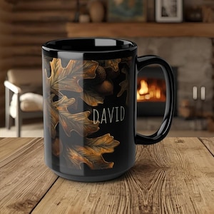 May include: Black ceramic mug featuring a design of autumn oak leaves and acorns in shades of brown and gold. The name "DAVID" is printed in white. The mug has a large handle.
