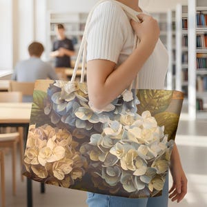 May include: A large tote bag featuring a floral design of blue and cream hydrangeas. The bag has a rectangular shape with a cream-colored rope handle. The floral pattern is detailed with green leaves and a vintage aesthetic.