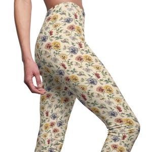 May include: Cream-colored leggings with a repeating floral pattern. The design features colorful pansies in shades of yellow, purple, and red, along with green leaves and stems. The leggings are form-fitting and made of a soft material.