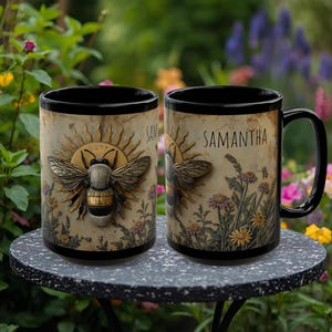 May include: Two black coffee mugs with a bee and sun design. The bee is in the center of the mug, with the name "Samantha" printed on the side. The mugs are sitting on a gray table with a garden in the background.