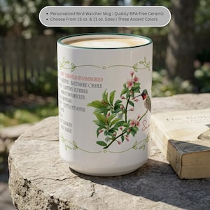Peut inclure: Un mug en céramique blanche avec un bord vert, orné d'une illustration botanique d'un colibri posé sur une branche fleurie. Le mug présente une liste de noms d'oiseaux en noir et le prénom Sam en rouge, avec un motif de bordure en vigne verte.