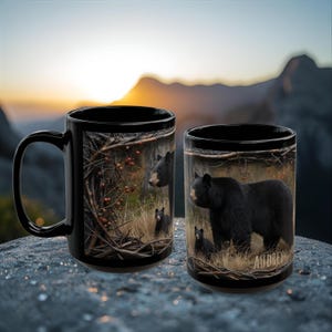 May include: Two black ceramic mugs with bear illustrations. One mug features a bear family in a forest setting, the other mug has a large bear with two cubs. The name "Audrey" is printed on the second mug.