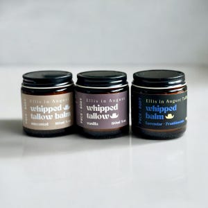 May include: Three amber glass jars with black lids and white labels. The labels read "Ellis in August" and feature a cowboy hat graphic. The jars contain whipped tallow balm in unscented, vanilla, and lavender-frankincense scents.
