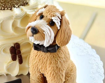 Puede incluir: Un adorno para pastel de perro de fondant marrón y blanco con una pajarita negra. El perro está sentado en un soporte para pastel plateado con un patrón de encaje.