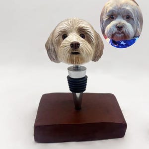 Puede incluir: Un tapón de botella con temática de perro con una escultura detallada de la cabeza de un perro. El tapón está montado sobre una base cuadrada marrón oscuro. Una imagen ovalada de un perro con un pañuelo azul está en el fondo.