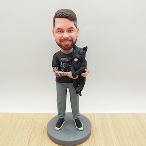 May include: A figurine of a person holding a black cat. The person is wearing a black t-shirt with the text "POWER" and tattoos on their arms, grey trousers, and black trainers. The cat has its tongue out.