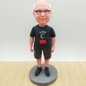 Może przedstawiać: Spersonalizowana figurka bobblehead przedstawiająca mężczyznę w okularach, czarnym t-shircie z logo "C" i brązowych szortach. Figurka stoi na szarej podstawie z napisem "DAVID STONE". Figurka ma na sobie czarne trampki.