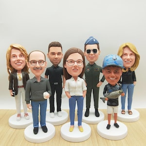May include: A group of custom bobblehead figures, each representing a person in different outfits and poses. The figurines are on white circular bases. The figures include a person in a military uniform, a person holding a fish, and others in casual wear.