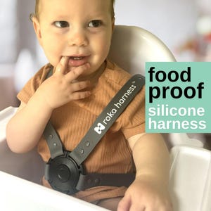 May include: A baby wearing a gray silicone harness with the text "roka harness" on the strap. The baby is wearing a brown shirt and sitting in a white high chair. The text "food proof silicone harness" is in the image.