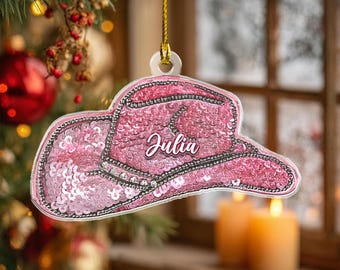May include: A pink sequined cowboy hat Christmas ornament with the name "Julia" in cursive script. The ornament is hanging from a gold string against a blurred background of a Christmas tree and candles.