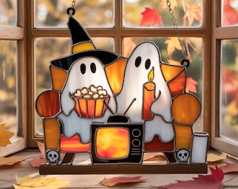 May include: Stained glass Halloween decoration featuring two ghosts, one wearing a witch hat, holding popcorn, and the other holding a candle. A retro TV sits between them. The design includes orange, white, black, and brown colors, with a skull on each side.