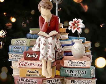 May include: A decorative ornament featuring a figure of a person reading atop a stack of books. The figure wears a red top and a floral patterned skirt. The books have titles like "Loved" and "Precious". A vase with a flower is also present.