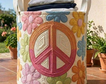 May include: A round, quilted laundry hamper with a peace sign and flower design. The hamper is cream-colored with a large peace symbol in the center, surrounded by colorful flowers. It has white handles and is filled with folded clothes.
