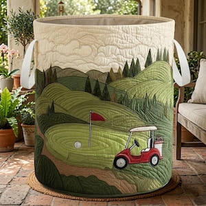 May include: Quilted storage bin with a golf course design. The bin features a green golf course scene with a red golf cart, a golf ball, and a red flag. The bin has white handles and a neutral top.